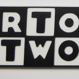 May include: A rectangular sign with the words "CARTOON NETWORK" in bold, black letters. The letters are set against a white background within black squares, creating a high-contrast design. The sign is likely a decorative item.