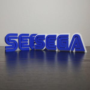 May include: A 3D-printed SEGA logo in blue and white. The letters are in a bold, stylized font, with the blue letters raised above the white base. The logo is displayed on a dark surface.