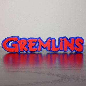 May include: A red and blue Gremlins logo, with the word "GREMLINS" in red, outlined in blue. The logo is placed on a dark surface, with a white background.