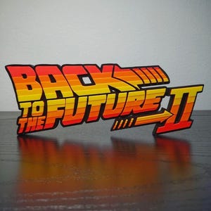 May include: A colorful, three-dimensional sign featuring the text "BACK TO THE FUTURE II" in bold, stylized lettering. The text is in orange, yellow, and red, with a black outline. The sign is placed on a dark surface.