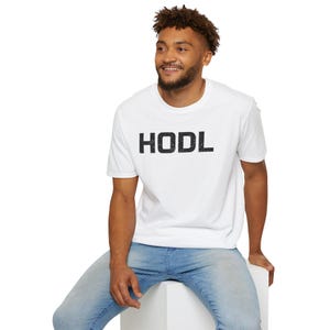 May include: White t-shirt with the word "HODL" printed in black. The person wearing the t-shirt is also wearing blue jeans and is sitting on a white surface. The t-shirt is short-sleeved.