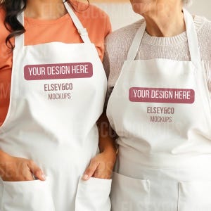 May include: Two white aprons with pockets and adjustable neck straps. Each apron has a rectangular maroon label that reads "YOUR DESIGN HERE" above the text "ELSEY&CO MOCKUPS". The aprons are being worn by two people.