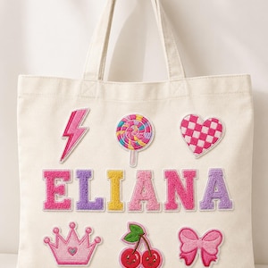 May include: A cream-colored canvas tote bag featuring the name ELIANA in large, multi-colored varsity-style chenille patches. The bag is decorated with embroidered patches of a pink lightning bolt, a lollipop, a checkered heart, a crown, cherries, and a bow.