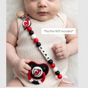 May include: A baby pacifier with a black, red, and white design. The pacifier clip features the name "NAME" spelled out with white letter beads. The clip also has red, black, and white beads. The text "*Pacifier NOT included*" is visible.