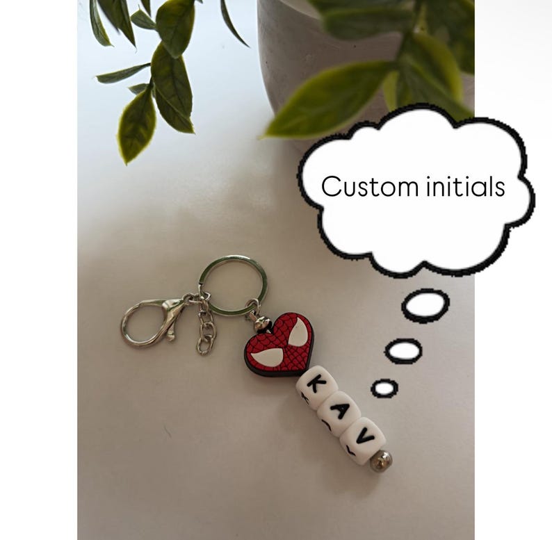 Puede incluir: Un llavero personalizado con el texto "Custom initials". El llavero presenta un dije en forma de coraz&oacute;n rojo con un dise&ntilde;o de telara&ntilde;a y cuentas de letras blancas que deletrean "KAV". Incluye un llavero y un cierre plateados.