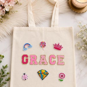 May include: A cream-colored canvas tote bag featuring the name GRACE in large, pink, glittery varsity-style letters. The bag is decorated with colorful patches including a rainbow, a lollipop, a pink crown, a ladybug, a diamond, and a flower.