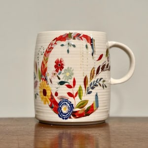 May include: Cream-colored ceramic mug with a floral design. The letter "C" is the focal point, adorned with colorful flowers and leaves. The mug has a ribbed texture and a curved handle, perfect for enjoying a warm beverage.