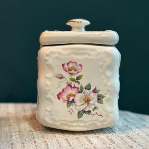 Op de afbeelding: Crèmekleurige keramische pot met deksel, met een bloemenontwerp van roze en witte bloemen met gele centra en groene bladeren. De pot heeft een textuur en een goudkleurige knop op het deksel.