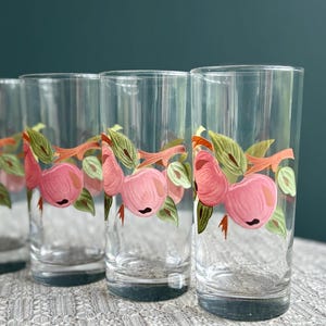 Puede incluir: Juego de cuatro vasos altos transparentes. Cada vaso presenta un diseño pintado a mano de melocotones rosas con hojas verdes y tallos naranjas. Los vasos están colocados sobre una superficie texturizada.