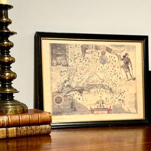 May include: Framed vintage map of Virginia with black frame. The map is in sepia tones with detailed illustrations and text, including the words "VIRGINIA" and "THE VIRGINIAN SEA". A stack of antique books and a brass candlestick are in the foreground.