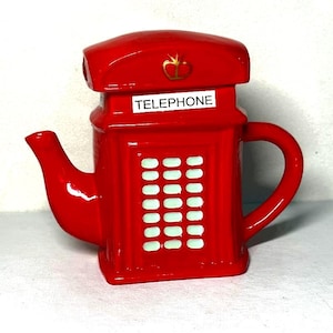 Könnte beinhalten: Eine leuchtend rote Keramik-Teekanne in Form einer klassischen britischen Telefonzelle. Die Teekanne verfügt über ein weißes Schild mit dem Wort TELEPHONE in schwarzen Buchstaben, ein goldenes Kronenemblem auf dem Dach und weiße Paneele.