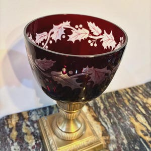 Puede incluir: Un cuenco de vidrio rojo rubí con un diseño de hojas y bayas de acebo blanco, montado sobre una base de pedestal de metal dorado. El borde del cuenco también está adornado con dorado. El cuenco probablemente se utiliza con fines decorativos.