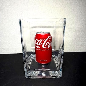 Puede incluir: Una lata roja de Coca-Cola dentro de un vaso transparente y cuadrado. La lata presenta el logotipo de Coca-Cola en blanco y las palabras "ORIGINAL TASTE" y "Delicious & Refreshing". El vaso tiene una base gruesa y lados rectos.