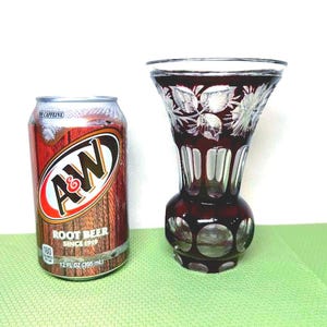 Op de afbeelding: A&W Root Beer-blikje naast een robijnrode glazen vaas met geëtste bloemmotieven. De vaas heeft een bolvormige basis en een uitlopende bovenkant. Het blikje is bruin met het A&W-logo en de tekst "ROOT BEER SINCE 1919".
