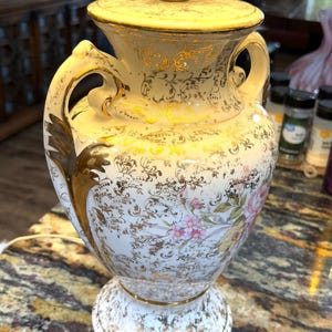 Könnte beinhalten: Eine Vintage-Tischlampe aus weißer Keramik mit goldenen Akzenten und floralen Designs. Die Lampe hat einen dekorativen urnenförmigen Sockel, zwei Griffe und einen goldfarbenen Lampenschirm. Die Lampe ist eine dekorative Leuchte.