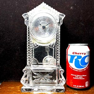 May include: A clear, decorative glass clock with a silver clock face and Roman numerals. The clock has a rectangular base and a peaked top. A red and white Cherry RC Cola can is next to the clock. The can is 12 fl oz (355 ml).