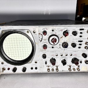 Op de afbeelding: Vintage oscilloscoop met een groot cirkelvormig scherm en talrijke wijzerplaten, knoppen en schakelaars. Het apparaat heeft een zilver en zwart kleurenschema, met labels en markeringen. Het scherm toont een rasterpatroon. Een klassiek elektronisch apparaat.