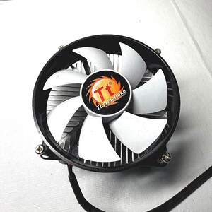 Puede incluir: Un ventilador de refrigeración de CPU de computadora circular con aspas blancas y un marco negro. El centro presenta un logotipo naranja y blanco con el texto "Tt Thermaltake". Un cable de alimentación negro se extiende desde la base del ventilador.