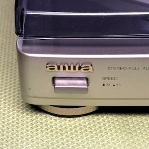 May include: Silver and black vintage Aiwa stereo turntable. The gold Aiwa logo is centered on the front. A silver switch is below the logo. The speed settings are 33 and 45. The turntable has a clear plastic cover.