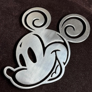 May include: A silver-toned metal cutout of a cartoon mouse head featuring large circular ears with spiral detailing, a wide smiling mouth, and oval eyes. The surface has a brushed metallic finish with clean, laser-cut edges and a flat profile.