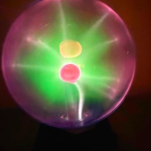 May include: A plasma ball with a clear glass exterior. The interior glows with green and pink light, with yellow and red orbs. The light emanates from the center, creating a captivating visual effect. The base is black.