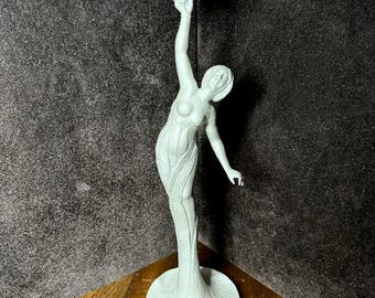 May include: A light green sculpture of a woman holding a flower aloft. The figure is slender, with flowing lines and a detailed floral base. The sculpture is made of a light green material and stands on a wooden surface.
