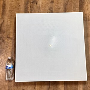 May include: A large, square, white perforated panel with a small, centered, rectangular accent. A clear plastic water bottle with a blue and white label is to the left of the panel. The panel is on a wooden surface.