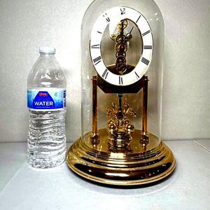 Puede incluir: Un reloj decorativo dorado con una esfera blanca y números romanos, encerrado en una cúpula de cristal transparente. Los intrincados mecanismos internos del reloj son visibles. La base es redonda y brillante. Una botella de agua está al lado del reloj.