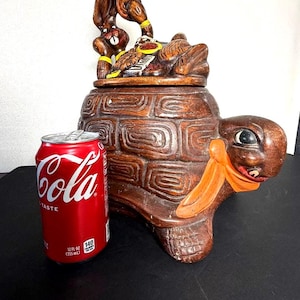 May include: A brown ceramic turtle-shaped cookie jar with a lid featuring a rabbit and turtle design. The turtle has an orange scarf. A red Coca-Cola can is next to the jar. The jar is a decorative kitchen item.