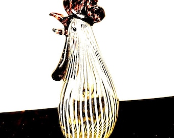 Op de afbeelding: Een glazen haanfiguur met een helder lichaam voorzien van verticale witte strepen, een donkerbruine kam en donkerbruine lellen. De haan is in profiel afgebeeld met de snavel open en heeft een glad, doorschijnend en reflecterend oppervlak.