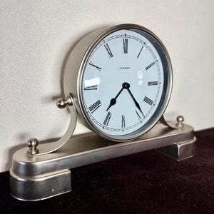 May include: A silver-toned desk clock with a white face and black Roman numerals. The clock has a curved base with decorative ball accents and the brand name "CHANEY" is printed on the clock face.