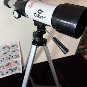 May include: A white and black Gskyer telescope mounted on a silver tripod. The telescope has a black eyepiece and a black lens cover. The Gskyer logo is printed on the white tube. Assembly instructions are visible in the background.