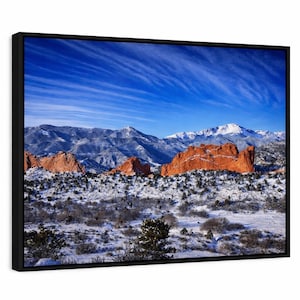 Puede incluir: Una impresión enmarcada de un paisaje invernal de montaña. La imagen muestra montañas cubiertas de nieve, formaciones rocosas rojas y un cielo azul brillante con nubes tenues. La obra de arte está encerrada en un marco negro.