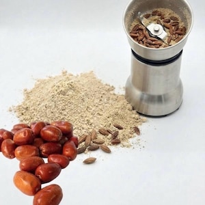 May include: A stainless steel electric spice grinder containing brown seeds, positioned next to a pile of light beige ground powder and a cluster of whole, oval-shaped reddish-brown jujube fruits on a plain white surface.