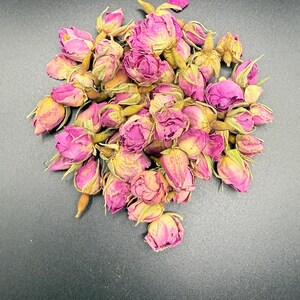 May include: A pile of dried rose buds, displaying a mix of pink and yellow colours. The buds are tightly closed, suggesting they are suitable for teas or potpourri. The background is a solid black.