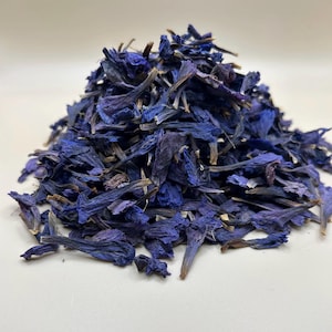 Puede incluir: Un montón de pétalos de aciano secos. Los pétalos son de color azul intenso con algunas puntas marrones, y están apilados sobre una superficie blanca. La imagen está bien iluminada y muestra la textura de los pétalos.