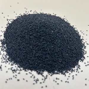 May include: A mound of small, dark blue seeds on a white surface. The seeds are tightly clustered, creating a dense pile. A few seeds are scattered around the edges of the main pile.