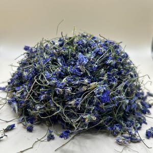 May include: A pile of dried blue delphinium flowers with green stems and leaves. The flowers are a vibrant blue color, and the stems are a mix of green and brown. The flowers are scattered on a white surface.