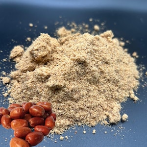 May include: A pile of light brown powder next to a cluster of reddish-brown, oval-shaped berries. The powder has a fine, granular texture. The berries are smooth and shiny. The background is a dark blue color.