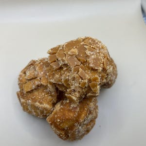 May include: A close-up shot of several pieces of brown, textured candy. The candy has a rough, uneven surface with a light-colored, crystalline coating. The pieces are clustered together, suggesting a snack or treat.