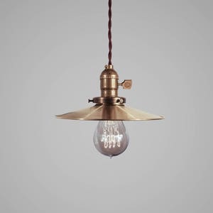 Könnte beinhalten: Eine Messing-Pendelleuchte mit einem breiten, flachen Schirm und einer klaren Glühbirne. Die Leuchte hängt an einem braunen Kabel.
