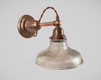May include: A vintage wall-mounted light fitting with a clear, ribbed glass shade and a copper-coloured metal fixture. The fixture has a circular wall mount, a curved arm, and a decorative bulb holder. The wiring is a twisted brown cord.