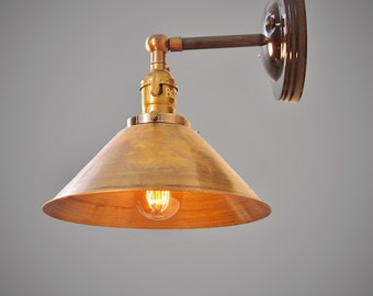 May include: A brass wall sconce with a cone-shaped shade and a black metal base. The light is on and the bulb is glowing.