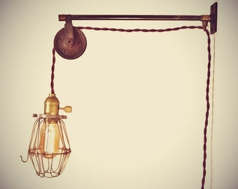 May include: A vintage industrial style pendant light with a caged bulb and a pulley system. The light is suspended from a metal arm attached to the wall. The cord is a dark red color.