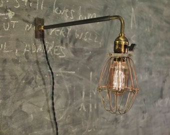 May include: A brass wall-mounted light fixture with a black wire cord and a caged light bulb. The light fixture has a long arm that extends out from the wall.