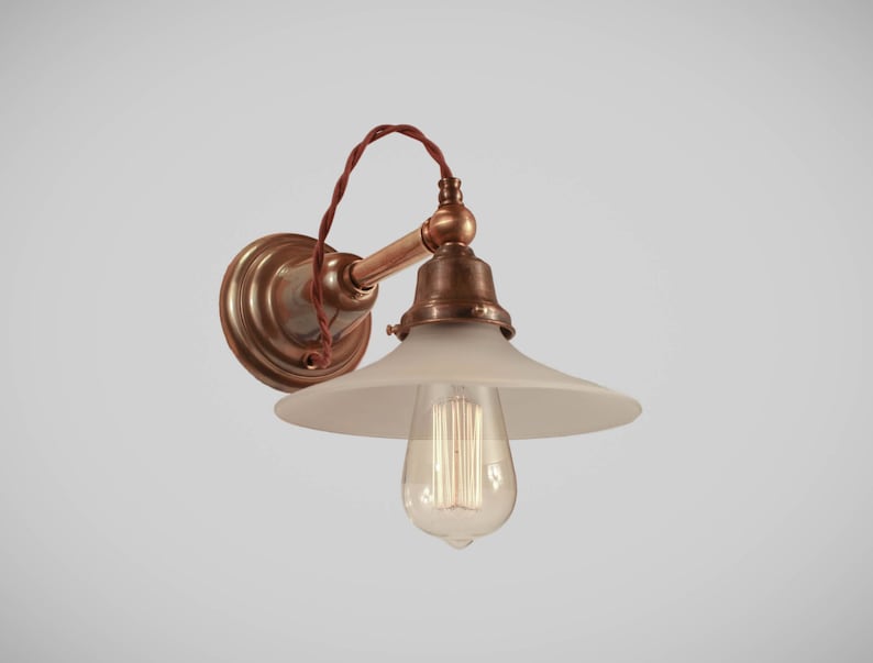 May include: An antique-style wall sconce with a frosted glass shade and exposed filament bulb. The fixture features a copper-colored arm and a round, brass-toned wall plate. A twisted brown wire adds a vintage touch.