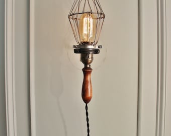 May include: A wall-mounted light fixture featuring a vintage-style Edison bulb enclosed in a wire cage. The base of the light is constructed from a polished wooden handle, connected to a black twisted fabric cord that hangs downward.