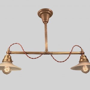 May include: A brass two-light pendant light fixture with a long, straight stem and two curved arms. Each arm has a white glass shade and a light bulb.