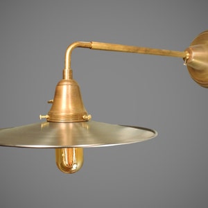 May include: A brass wall sconce with a silver metal shade and a single light bulb. The sconce has a curved arm and a round base.