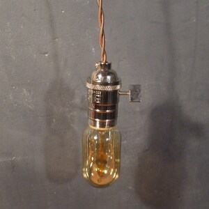 May include: A close-up of a hanging light bulb with a brown braided cord and a chrome socket. The light bulb is clear glass and has a warm yellow glow.